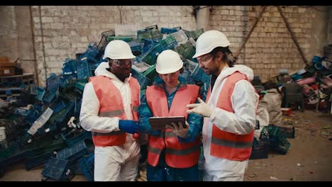 A-trio-of-waste-recycling-plant-workers-talk-about-their-plans-and-look-at-a-tablet-screen-while-standing-near-a-large-pile-of-plastic-waste-at-the-plant