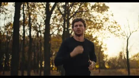 Un-Hombre-Seguro-De-Sí-Mismo,-Con-Cabello-Rizado-Y-Barba,-Con-Un-Uniforme-Deportivo-Negro,-Corre-Por-Un-Sendero-Forestal-En-Un-Bosque-Otoñal-Con-Hojas-Marrones-Caídas-Al-Atardecer.