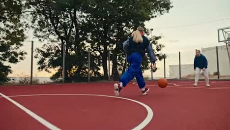 Filmada-Desde-Abajo,-Una-Chica-Rubia-Con-Uniforme-Deportivo-Corre-Y-Supera-A-Todos-Los-Oponentes,-Lanzando-Una-Pelota-De-Baloncesto-Naranja-Al-Aro-En-Una-Cancha-Callejera-Roja-Por-La-Mañana.