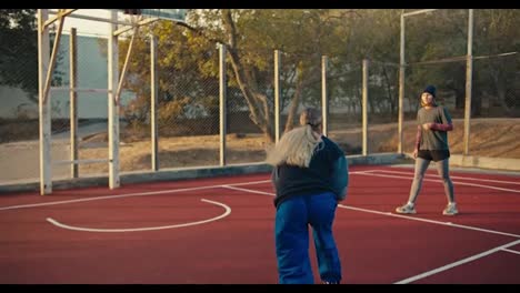 A-blonde-girl-in-a-sports-uniform-gives-a-pass-with-an-orange-sword-to-her-friend-who-throws-the-ball-into-the-basket-during-their-game-of-basketball-on-a-red-street-court-in-the-morning