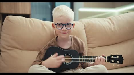 Portrait-of-a-happy-little-albino-boy-with-white-hair-color-in-blue-round-glasses-who-plays-a-black-ukulele-and-sits-on-a-cream-sofa-in-a-modern-apartment-at-home