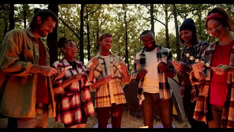 A-happy-company-of-six-people-in-special-camping-clothes-in-plaid-shirts-stand-around-a-fire-and-string-marshmallows-onto-small-wooden-sticks-in-a-green-autumn-forest.-The-fire-illuminates-the-hikers-with-red-orange-color-during-the-evening