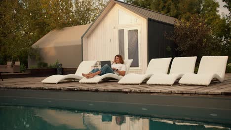 A-curly-haired-guy-in-glasses-is-lying-on-a-white-deckchair-near-the-sunbeds-by-the-pool-and-working-on-a-laptop.-Rest-in-the-country-house