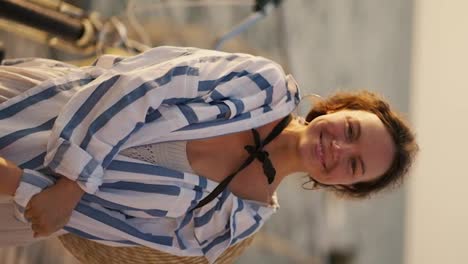 Vertical-video-portrait-of-a-brown-haired-girl-with-large-earrings-who-is-dressed-in-a-white-blue-shirt-and-a-black-bow.-Happy-brown-haired-girl-smiles-and-looks-at-the-camera-against-of-the-sea