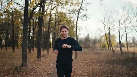 A-confident-and-slightly-tired-happy-man-with-curly-hair-and-a-beard-in-a-black-sports-uniform-looks-at-his-watch-during-his-morning-jog-in-the-autumn-sunny-forest