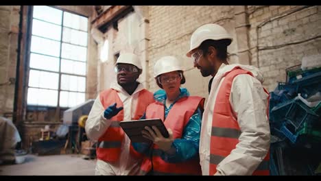 Shooting-close-up-a-brunette-man-in-a-white-uniform-a-brunette-girl-in-a-White-helmet-and-with-a-tablet-in-her-hands-and-their-employee-with-Black-skin-in-a-white-special-uniform-sorting-out-their-plans-and-discussing-work-at-a-waste-processing-plant-near-a-large-pile-of-plastic-garbage