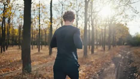 Rückansicht-Eines-Mannes-Mit-Lockigem-Haar-In-Einer-Schwarzen-Sportuniform,-Der-Beim-Morgendlichen-Joggen-In-Einem-Herbstmorgenwald-Bei-Strahlender-Sonne-Auf-Einem-Erdweg-Läuft