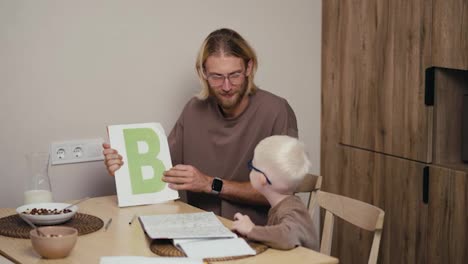 Ein-Blonder-Mann-Mit-Brille-Und-Bart-Zeigt-Seinem-Kleinen-Albino-Sohn-Mit-Blauer-Brille-Buchstaben-Auf-Englisch-Und-Bringt-Ihm-Grammatik-Auf-Englisch-Bei,-Während-Er-In-Der-Küche-Seine-Hausaufgaben-In-Der-Grundschule-Vorbereitet