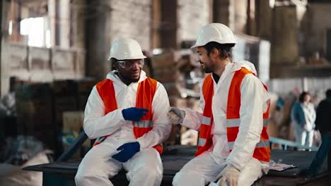 A-man-with-Black-skin-in-a-white-protective-uniform-and-an-orange-vest-plays-rock-paper-scissors-with-a-man-and-loses-to-him-during-his-fun-and-break-at-work-at-a-waste-recycling-and-sorting-plant