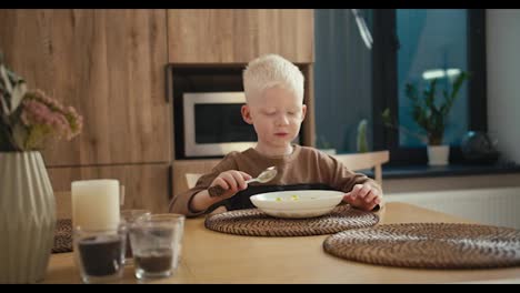 Ein-Glücklicher-Kleiner-Albino-Junge-Mit-Weißer-Haarfarbe-Und-Blauen-Augen-Isst-Sein-Frühstück-Am-Morgen,-Während-Er-In-Einer-Modernen-Apartmentküche-Am-Tisch-Sitzt.-Ein-Glücklicher-Junge-Isst-Sein-Frühstück-Mit-Einem-Löffel-Von-Einem-Teller,-Während-Er-Auf-Einem-Weißen-Stuhl-Am-Tisch-Sitzt