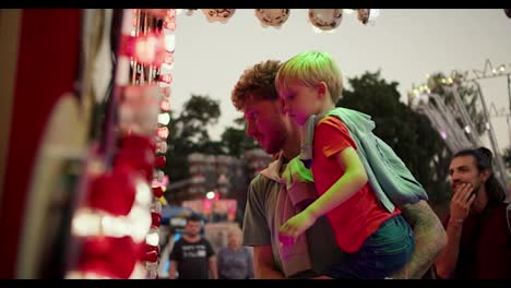 A-dad-with-curly-hair-in-a-Green-T-shirt-holds-his-little-blond-son-in-a-red-T-shirt-in-his-arms-and-they-stand-near-the-ticket-office-in-an-amusement-park-that-is-equipped-with-bright-light-bulbs