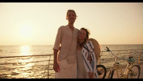 Portrait-of-a-happy-couple,-a-guy-in-light-clothes-and-a-girl-in-a-blue-white-shirt-with-a-Straw-hat-on-the-back,-hugging-and-looking-at-the-camera-against-the-backdrop-of-their-bicycles-on-the-beach,-which-is-covered-with-boards-near-the-sea-at-Sunrise-in-summer