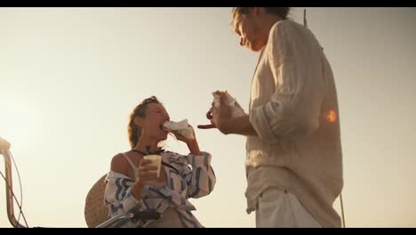 Shot-from-below-against-the-sky,-a-guy-and-a-girl-in-light-beach-clothes-eat-hot-dogs-and-drink-coffee-near-the-sea-in-the-summer.-A-guy-straightens-a-girl's-hair-during-a-date-at-sea