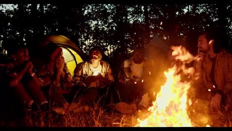 Evening-cozy-communication-near-the-fire,-a-small-group-sits-near-the-fire-and-tells-scary-stories-against-the-backdrop-of-the-floor-and-the-evening-autumn-forest.-A-man-in-a-hat-shines-a-flashlight-in-his-face-and-tells-scary-stories-to-a-group-during-a-hike