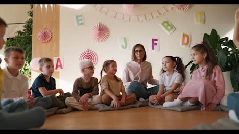 Ein-Glückliches-Blondes-Mädchen-Mit-Einer-Bob-Frisur,-Einer-Brille-Und-Einem-Weißen-Hemd-Sitzt-Mit-Ihren-Schülern-Auf-Einem-Speziellen-Kissen-Auf-Dem-Boden-Im-Clubraum-Zur-Vorbereitung-Der-Kinder-Auf-Die-Schule-Und-Kommuniziert-Mit-Ihnen