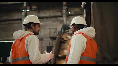 A-man-with-a-beard-in-a-white-helmet-and-a-white-protective-uniform-stands-and-communicates-with-his-colleague-a-man-with-Black-skin-in-an-orange-vest-near-a-conveyor-belt-at-a-large-gray-waste-recycling-plant