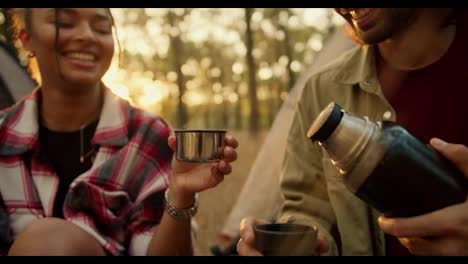 Close-up-shot-of-a-brunette-man-in-a-light-green-jacket-pouring-tea-from-a-thermos-of-a-girl-with-Black-skin-in-a-checkered-shirt-during-their-hike-in-a-light-green-sunny-summer-forest