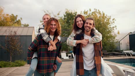 Portrait:-two-girls-are-sitting-on-the-backs-of-their-boyfriends-in-checkered-shirts-and-smiling-near-sunbeds.-Rest-in-the-country-house