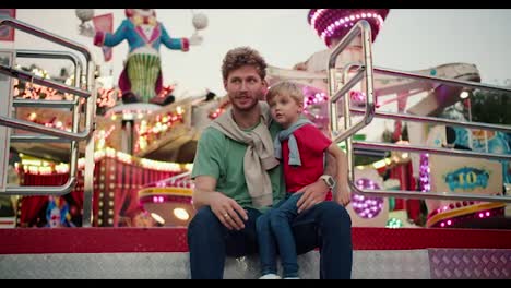 A-father-with-curly-hair-with-stubble-in-a-Green-T-shirt-sits-near-his-little-blond-son-in-a-red-T-shirt-who-sits-on-his-leg-and-looks-at-the-attractions-in-an-amusement-park-with-bright-lights