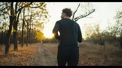 Rear-view-of-a-sporty-guy-in-a-black-sports-uniform-runs-along-an-earthen-path-in-the-autumn-forest-at-sunrise-in-the-morning