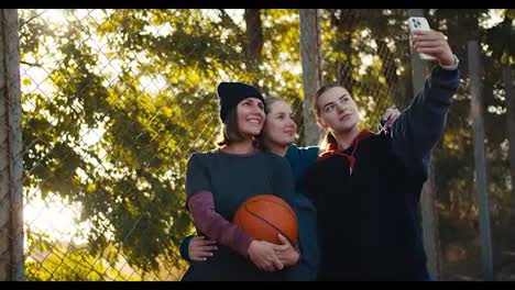 A-trio-of-happy-girls-basketball-players-in-sportswear-take-a-selfie-together-with-a-basketball-ball-on-an-outdoor-basketball-court-near-a-grating-on-a-sunny-day