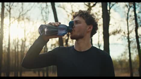 Confident-male-athlete-with-curly-hair-drinks-water-from-a-special-gray-sports-bottle-while-resting-after-a-run-in-the-autumn-forest-on-a-sunny-day.-A-man-in-a-black-sports-uniform-drinks-water-and-rests-after-playing-sports-and-jogging