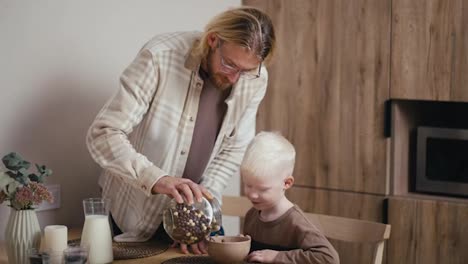 Happy-blond-man-in-glasses-with-a-beard-pours-cereals-of-different-colors-to-his-little-albino-son-with-white-hair-color-in-a-bowl-and-then-adds-milk-and-makes-breakfast-in-the-morning-for-his-little-son-in-a-modern-kitchen