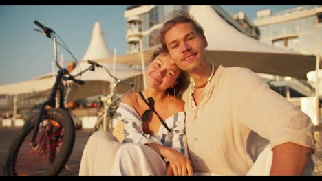 Portrait-of-a-couple:-A-girl-in-light-clothes-leans-on-the-shoulder-of-a-guy-with-stubble-in-a-white-beach-shirt-against-the-background-of-white-cafe-umbrellas.-A-guy-and-a-girl-rode-a-bicycle-to-the-beach