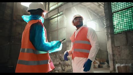 Two-colleagues-in-special-uniforms-and-orange-vests-communicate-at-a-large-gray-waste-recycling-plant.-A-man-and-a-girl-communicate-at-a-waste-processing-plant