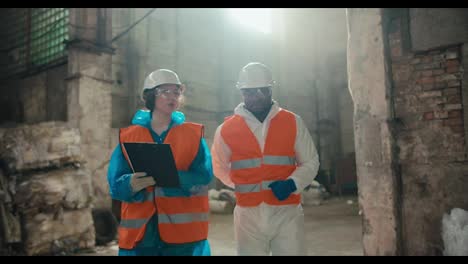 Un-Dúo-De-Ingenieros,-Una-Chica-Con-Ropa-Protectora-Y-Un-Chaleco-Naranja,-Camina-Con-Un-Chico-De-Piel-Negra-Y-Casco-Blanco-A-Través-De-Una-Enorme-Y-Antigua-Planta-Procesadora-De-Residuos-Y-Se-Comunican-Mientras-Sostienen-Una-Tableta-En-Sus-Manos.