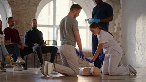 Professional-nurse-in-white-medical-uniform-teaches-bearded-brunette-to-do-artificial-respiration-mannequin-on-a-practical-course-for-a-medic