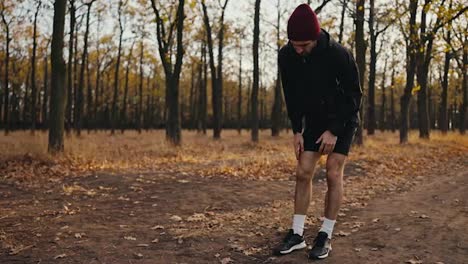 Ein-Brünetter-Mann-In-Schwarzer-Sportuniform-Und-Roter-Mütze-Streckt-Sein-Verletztes-Bein,-Während-Er-Morgens-Im-Herbstlichen-Wald-Auf-Einem-Erdweg-Joggt