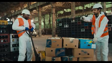 A-Black-man-in-a-white-uniform-together-with-his-colleague-a-man-with-a-beard-and-an-orange-vest-are-trying-to-put-a-wheelbarrow-with-sorted-waste-in-place-near-boxes-with-sorted-glass-waste-at-a-waste-recycling-plant