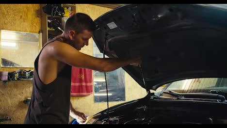 A-male-master-motorist-mechanic-with-a-short-haircut-in-a-gray-T-shirt-checks-the-hood-of-the-car-and-the-oil-there-in-his-garage-workshop.-Car-hood-repair-in-my-workshop