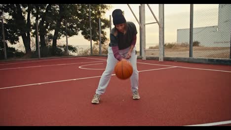 Portrait-of-a-girl-with-a-bob-hairstyle-in-a-black-hat-who-bounces-an-orange-basketball-off-the-floor-on-a-basketball-court-and-then-looks-at-the-camera-and-poses-in-the-morning-during-her-basketball-training