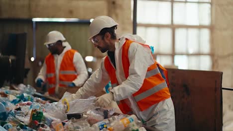 A-brunette-man-with-a-beard-in-a-white-protective-uniform-and-an-orange-vest-together-with-his-colleagues-sorts-plastic-bottles-by-color-at-a-waste-recycling-plant