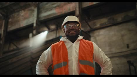 Retrato-En-Primer-Plano-De-Un-Hombre-Feliz-De-Piel-Negra-Con-Gafas-Y-Un-Casco-Blanco-Con-Un-Uniforme-Blanco-Especial-Y-Un-Chaleco-Naranja-Que-Se-Encuentra-En-Una-Enorme-Fábrica-Cerca-De-La-Cinta-Transportadora.