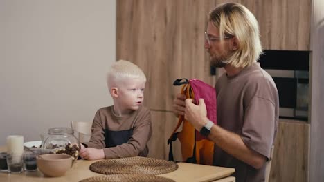 A-happy-albino-boy-with-white-hair-puts-on-a-bright-backpack-before-going-to-school-in-the-kitchen-and-his-blond-dad-with-a-beard-and-glasses-helps-him-with-this