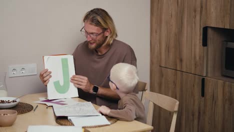 Ein-Blonder-Mann-Mit-Brille-Und-Bart-Zeigt-Seinem-Kleinen-Sohn,-Einem-Albino-Sohn-Mit-Brille-Und-Weißem-Haar,-Verschiedene-Buchstaben-Auf-Englisch-Und-Der-Junge-Benennt-Sie,-Während-Er-Zu-Hause-In-Einer-Modernen-Küche-Seine-Hausaufgaben-Macht