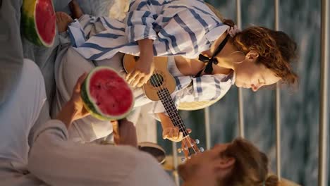 Vertical-video-of-a-happy-couple-on-a-date.-On-the-pier-near-the-sea,-the-guy-cuts-a-watermelon,-the-girl-plays-the-ukulele.-Guy-and-girl-on-a-picnic-on-the-pier-near-the-sea