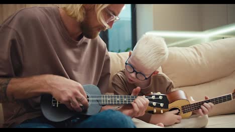 Ein-Kleiner-Albinojunge-Mit-Weißem-Haar-Und-Blauer-Brille-Sieht-Seinem-Vater-Beim-Gitarre--Und-Ukulelespielen-Zu-Und-Versucht,-Ihm-Nachzusprechen,-Während-Er-Auf-Dem-Sofa-In-Einer-Modernen-Wohnung-Sitzt.-Ein-Albinojunge-Lernt-Zu-Hause-Mit-Seinem-Vater-Ein-Musikinstrument-Zu-Spielen