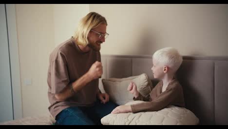 Feliz-Padre-Rubio-Con-Barba-Y-Gafas-Jugando-Con-Su-Pequeño-Hijo-A-Piedra,-Papel-O-Tijera-Y-Divirtiéndose-En-Una-Cama-Color-Crema-Cerca-De-Una-Computadora-Portátil-Gris-Durante-Sus-Vacaciones-De-Fin-De-Semana