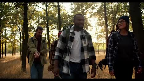 Happy-international-group-on-a-hike-in-the-summer-forest.-A-man-with-Black-skin-in-a-checkered-shirt-and-a-girl-with-a-bob-with-blond-hair-walk-through-the-forest-with-their-friends-in-special-hiking-clothes-in-the-summer