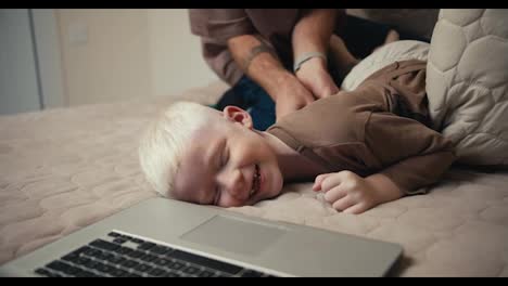 Close-up-shot-of-a-small-albino-boy-with-white-hair-in-a-brown-sweater-lying-on-the-bed-while-his-father-tickles-him-lying-near-the-laptop.-Happy-fun-time-for-parents-and-children-during-vacation-and-weekend