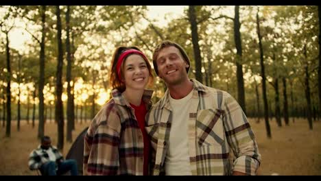 Portrait-of-a-happy-couple,-a-blond-man-with-stubble-in-a-plaid-shirt-and-a-brunette-girl-in-a-red-bandana-and-standing-and-looking-at-the-camera-leaning-on-each-other.-Happy-couple-during-a-hike-in-a-sunny-summer-green-forest