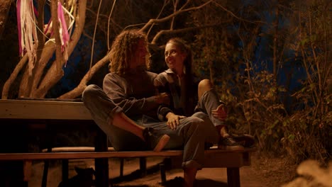 A-couple-in-love-is-sitting-on-a-wooden-bench-near-a-fire-and-talking.-Party-outside-the-city.-Rest-in-the-country-house