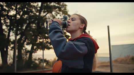 Primer-Plano-De-Una-Chica-Rubia-Con-Una-Sudadera-Con-Capucha-Negra-Bebiendo-Agua-De-Una-Botella-Deportiva-Gris-Mientras-Sostiene-Una-Pelota-De-Baloncesto-En-Su-Mano-Derecha-Durante-Su-Entrenamiento-Matutino-En-La-Cancha-De-Baloncesto-En-El-Verano.