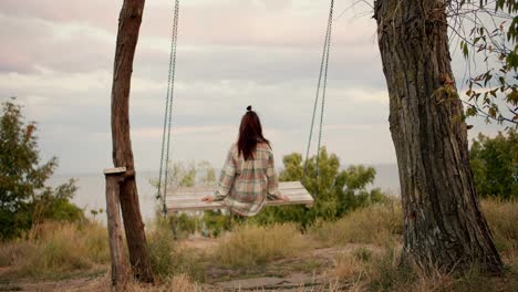 Shooting-from-behind:-A-brunette-girl-in-a-checkered-shirt-swings-on-a-wooden-swing-outside-the-city.-Rest-in-the-country-by-the-sea