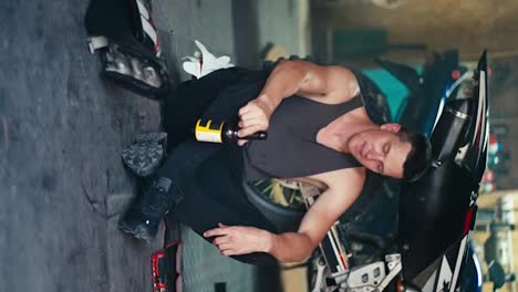 Vertical-video:-a-male-mechanic-with-a-short-haircut-in-a-gray-shirt-sits-on-the-floor-drinking-a-drink-from-a-brown-glass-bottle-in-a-workshop-garage