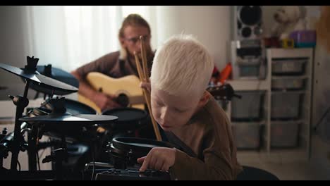 An-albino-boy-with-white-hair-sets-up-electric-drums-to-start-playing-his-favorite-tunes-along-with-his-father,-a-blond-man-in-glasses-with-a-beard.-Happy-before-musicians,-a-blond-man-with-a-beard-plays-an-acoustic-guitar-and-his-little-son-plays-along-with-him-on-the-drums